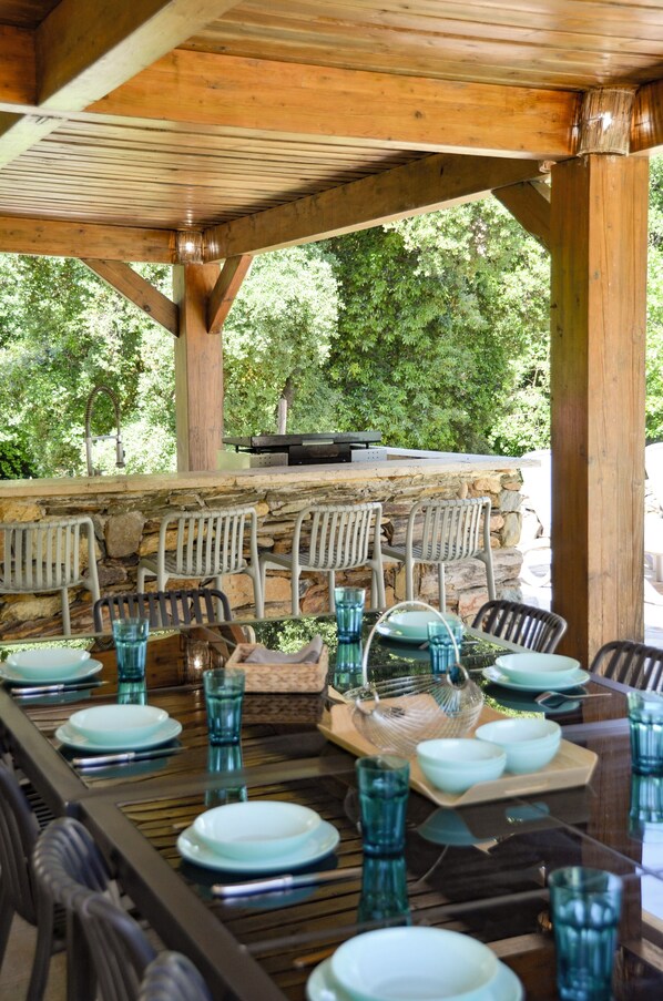 Un espace extérieur aménagé pour les repas, avec un mur en pierre, des poutres en bois et des chaises en métal.
