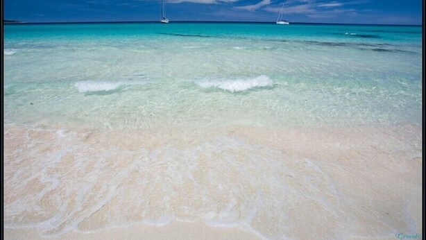 Een strand met helder turquoise water en twee boten in de verte.