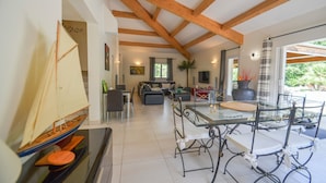 Een ruime woonkamer met een hoge plafond en zichtbare houten balken, een glazen eettafel met stoelen, een bank en een klein model zeilboot.