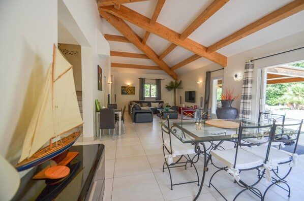 A spacious living area with a high ceiling and exposed wooden beams, a glass dining table with chairs, a sofa, and a small sailboat model on display. - Modern Villa With Heated Pool - Saint Florent - (Saint-Florent)
