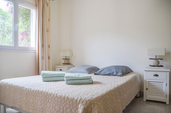 A neatly made bed with a quilted bedspread, two folded towels, and two patterned pillows. There is a small bedside table with a lamp and a decorative item on it.