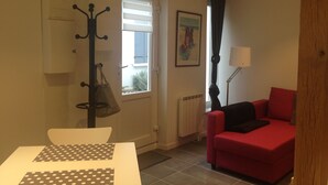 A room with a red sofa, a white table with a polka dot tablecloth, a coat rack with a bag, a window with blinds, a radiator, and a floor lamp.