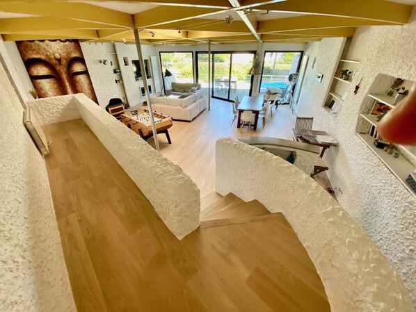 A spacious living area with a wooden floor, a sofa, a coffee table, and a foosball table. There is a large window providing a view of the outdoors.