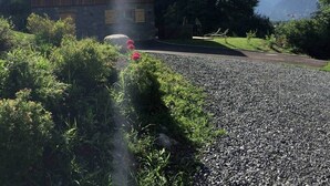 A rustic building with a stone facade, a gravel driveway, and a lush green landscape with trees and a mountain in the background.