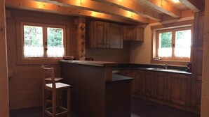 A wooden kitchen with a bar area, two windows, and a tiled floor.