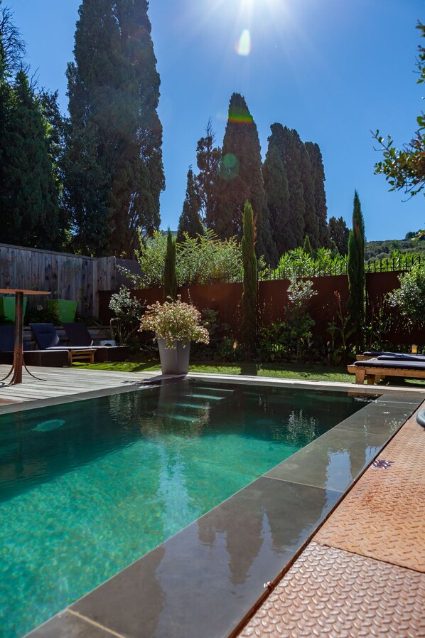 A clear swimming pool with a wooden deck and lounge chairs, surrounded by tall trees and a wooden fence.