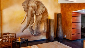 Una habitación con un gran mural de elefantes en la pared, una silla de madera, una mesita con una taza, y un mueble de madera grande.