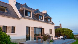 Ein modernes Haus mit geneigtem Dach, großen Fenstern und einer überdachten Terrasse mit Gartenmöbeln.