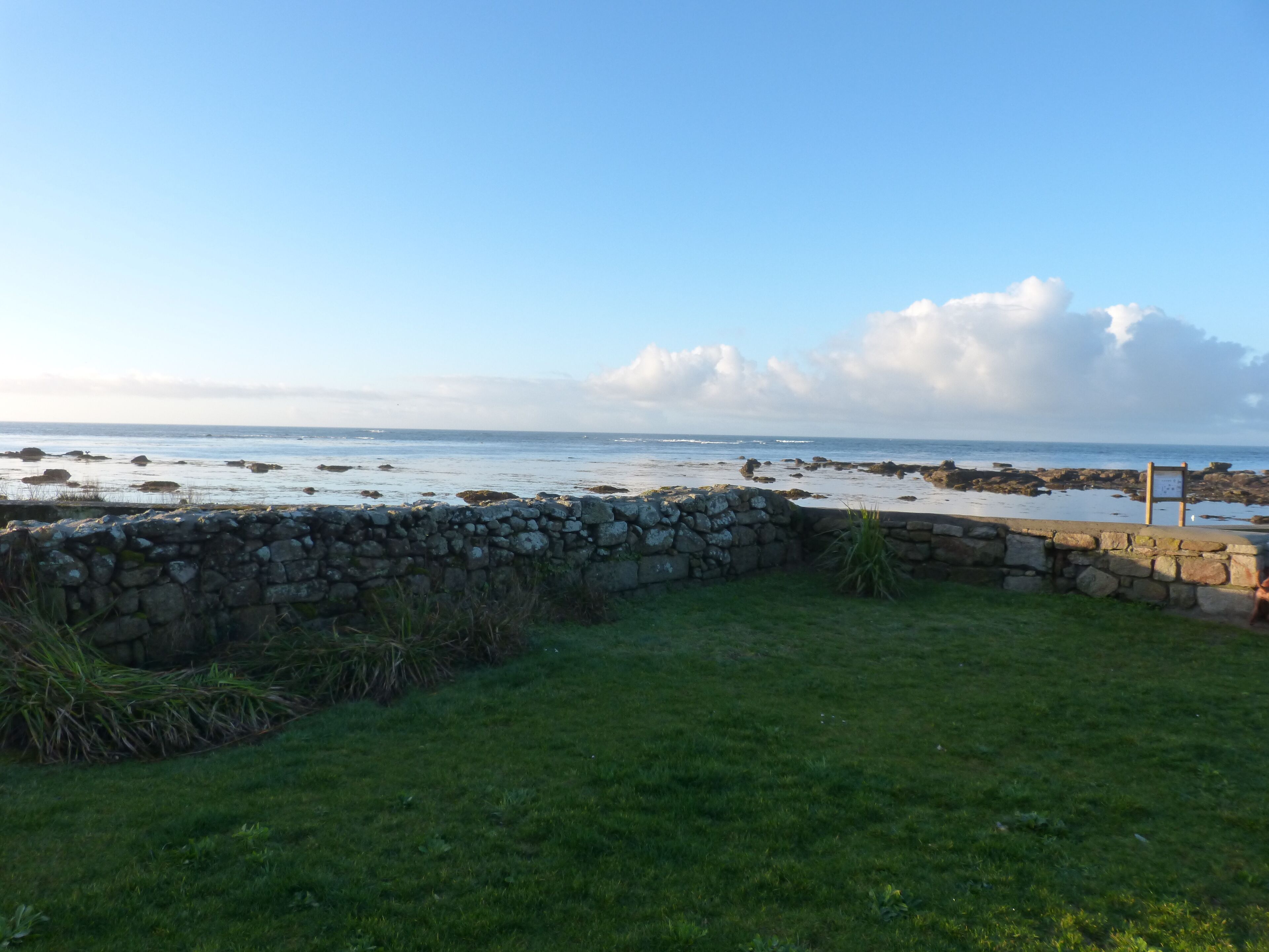 A coastal view with a stone wall, grassy area, and a signboard near the water.