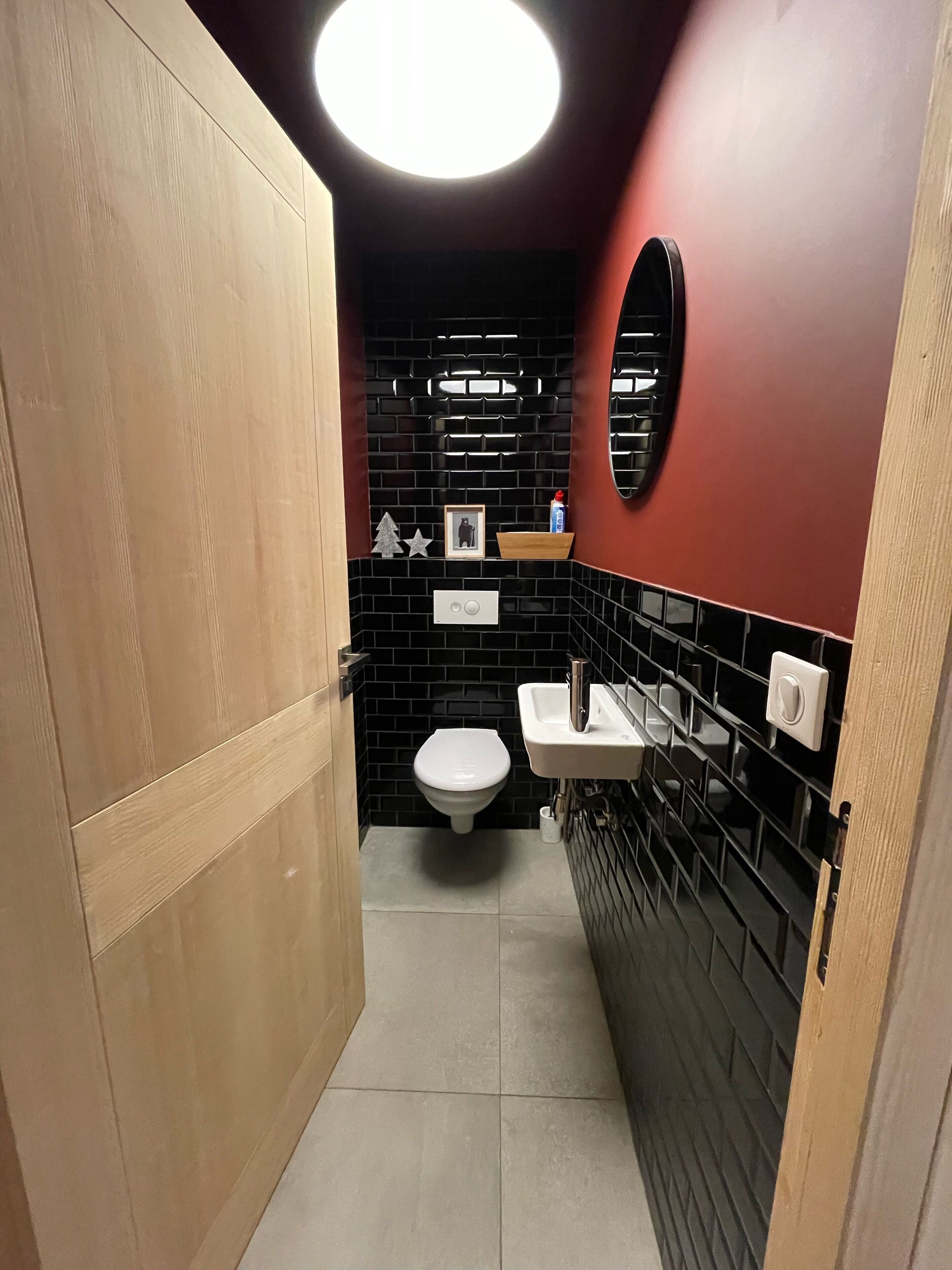 Ein kleines Badezimmer mit Toilette, schwarzen Fliesen, einer Holztür und einem weißen Waschbecken mit Spiegel darüber.