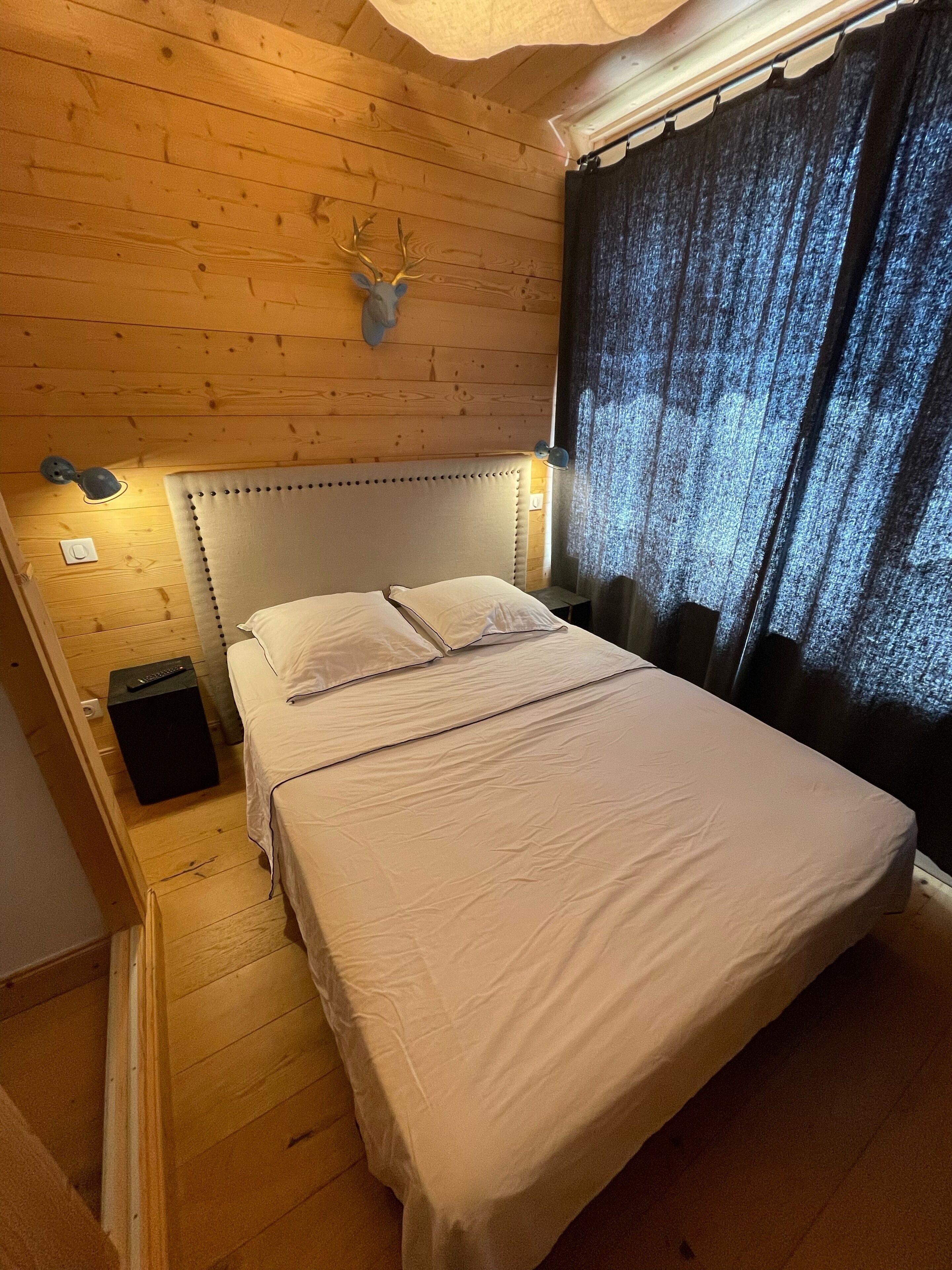 Een kamer met houten lambrisering, een bed, een opgehangen hertenkop en een gordijn.