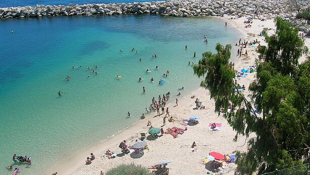 Una spiaggia con acque cristalline turchesi, una frangiflutti rocciosa e persone che nuotano e si rilassano sulla sabbia.