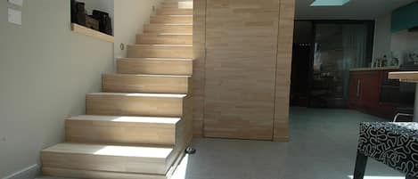Una escalera con escalones de madera que lleva a una plataforma, una cocina con armarios y electrodomésticos, y una mesa con estampado.