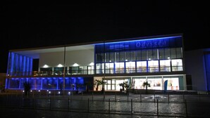 Un edificio moderno con un letrero que dice "Odyssea". Cuenta con amplios ventanales, techo plano y una zona pavimentada en su parte frontal.