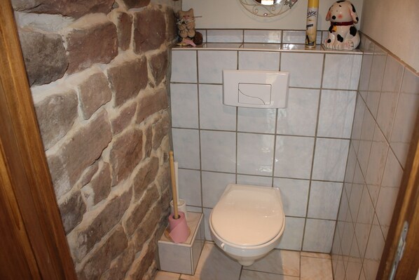 Une salle de bain avec un mur en pierre, des toilettes blanches, un sèche-mains et une petite étagère ornée d’objets décoratifs.