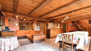 Un intérieur chaleureux et rustique, avec un plafond en bois, un canapé, une table à manger recouverte d’une nappe à motifs floraux, et un coin cuisine doté d’un mur en pierre et de meubles en bois.