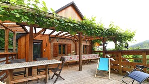 Une terrasse en bois avec une table et des chaises, un espace couvert également équipé d’une table et de chaises, et une vue sur les montagnes.