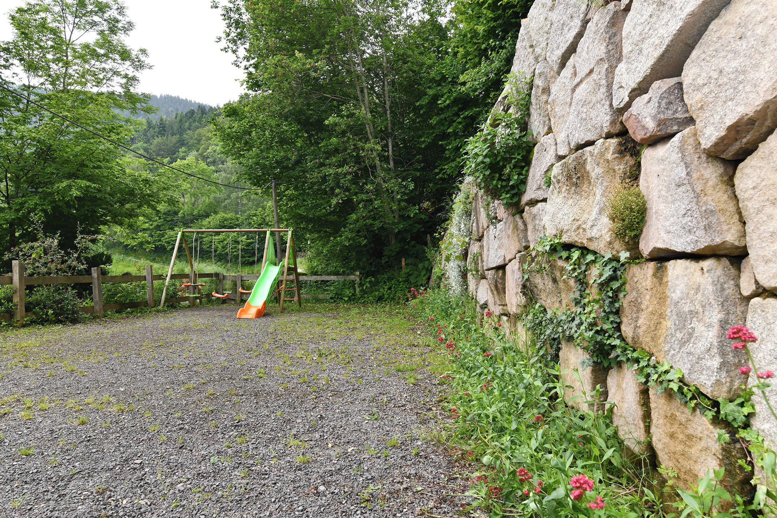 Ein Kiesweg führt zu einer hölzernen Schaukelanlage inmitten von Natur, mit einer Steinmauer und viel Grün.