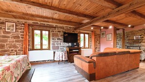 Une pièce chaleureuse avec un mur en pierre, un plafond en bois, un canapé, une télévision et une table recouverte d’une nappe à motifs floraux.
