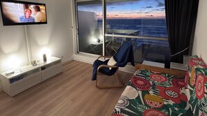 A hotel room with a TV, a bed with a colorful bedspread, a chair, a desk, and a large window with a view of the ocean. - Apart 4 pers. with swimming pool and unbelievable view on ocean ! (Biarritz)