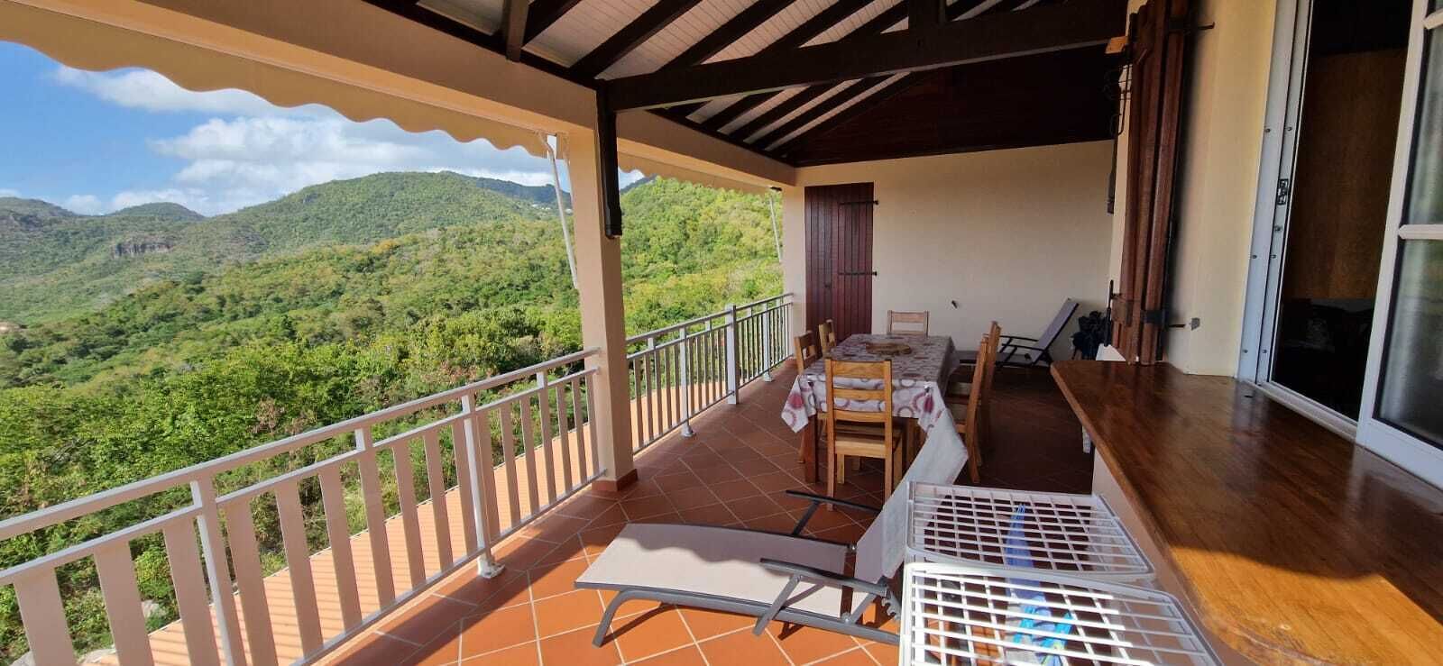 A balcony with a table and chairs, a view of green hills, and a wooden floor.
