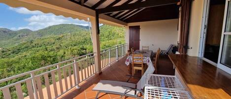 A balcony with a table and chairs, a view of green hills, and a wooden floor.