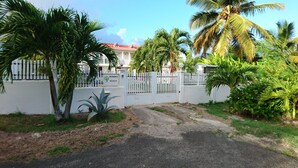 A gated entrance with a white fence, palm trees, and a building with a red roof in the background.