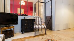 A living room with a large flat-screen TV on a wooden stand, a striped armchair, a dark wooden cabinet with drawers, and a large window with a view of a red wall and a hanging light fixture. - The Loisy - A very smart spot in the heart of Dijon for up to 4 people (Dijon)