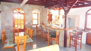 A cozy kitchen with a stone wall, wooden ceiling, and a dining area with wooden chairs and a glass table.