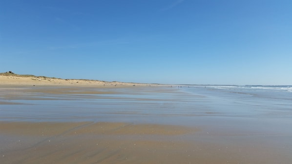 Una playa amplia de arena con olas suaves, cielo despejado y dunas al fondo.