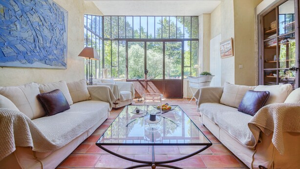 A spacious living area with a glass coffee table, two beige sofas with cushions, a large abstract painting on the wall, and a view through large windows to the outdoors. - Farmhouse in Provence, farmhouse, private in a 7 hectares park, heated pool (L'isle-Sur-La-Sorgue)