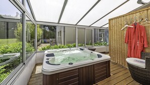A hot tub in a covered outdoor area with wooden flooring, a wooden wall, and a view of a garden.