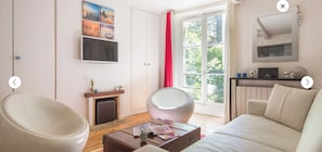 Un salon moderne comprenant un canapé, deux fauteuils, une cheminée, un téléviseur et une grande fenêtre ornée d’un rideau rouge.