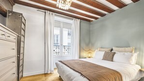 A bedroom with a wooden floor, a bed with white bedding and a brown throw, a dresser, and a window with white curtains. - Bahia, 2BR/1BA, 7 people (París)