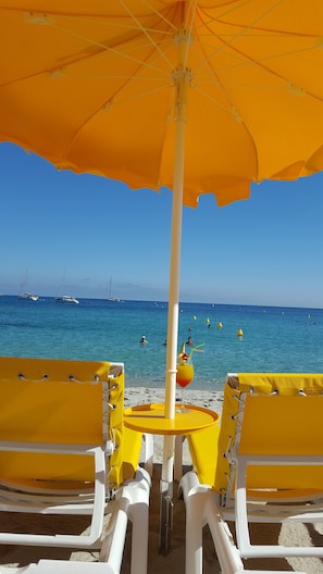 Ein Strandbild mit gelben Stühlen und einem Sonnenschirm, klares blaues Meer und Boote im Hintergrund.