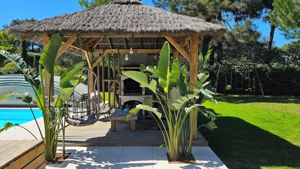 Um gazebo de madeira com telhado de palha, cercado por plantas tropicais, próximo a uma piscina e área de churrasco.
