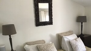 A bedroom with two beds, each with a white bedspread and patterned pillows, flanked by dark nightstands with lamps. A large ornate mirror hangs above the beds.