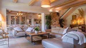 A spacious living room with a large sectional sofa, a coffee table, and a dining area with white chairs. There is a staircase leading to an upper level, and the room features exposed wooden beams and a stone accent wall. - In the Mas des Pampres, Provence is at your fingertips (Cabannes)