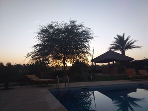 Una piscina con sillas de descanso y un pabellón, rodeado de árboles y con un atardecer de fondo.