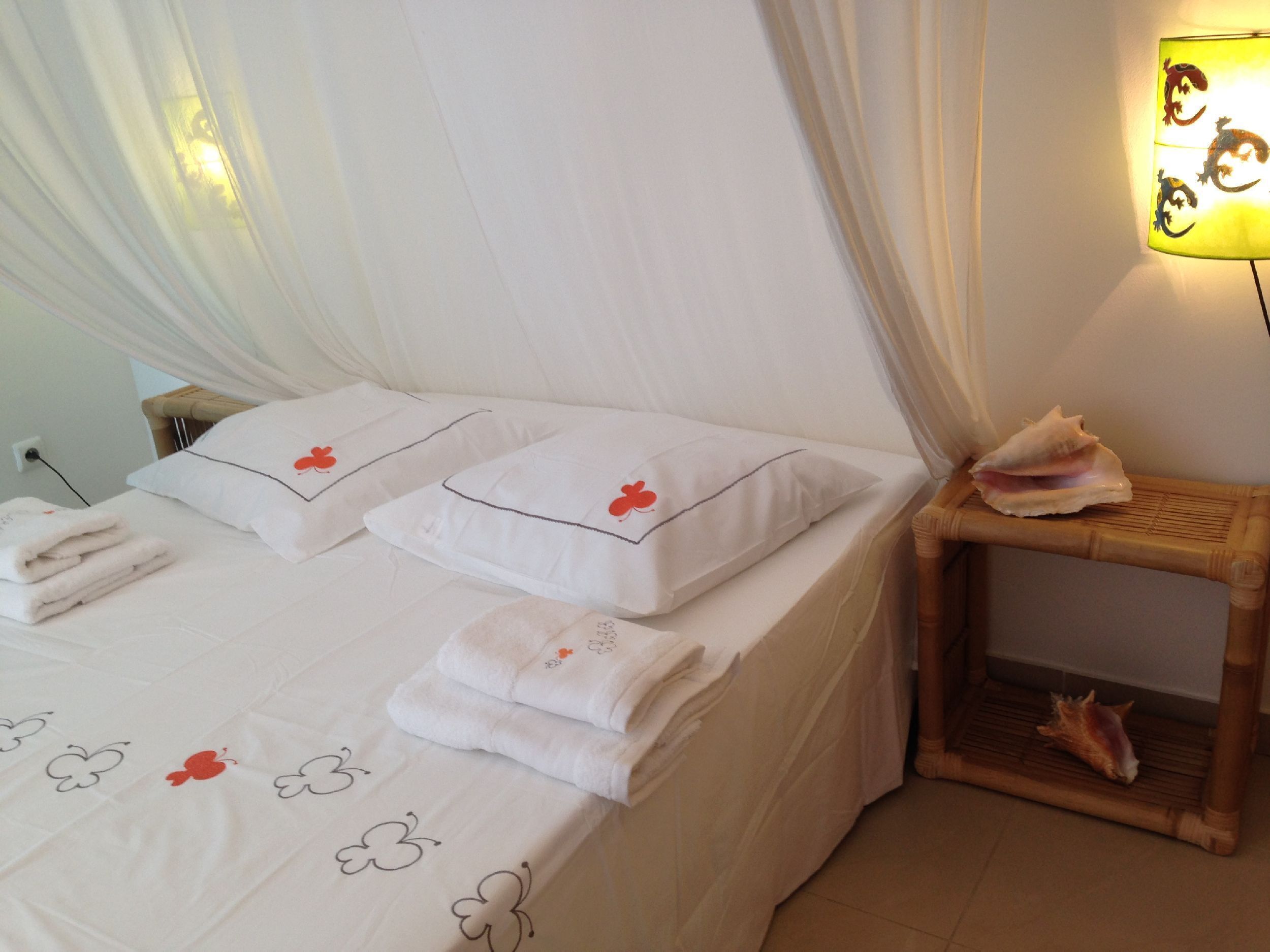 A bed with white linens and red floral pillows, a bedside lamp, and a small wooden side table with a shell.