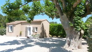Un petit bâtiment d’un seul étage, doté d’un toit en tuiles et entouré de gravier. Il y a deux fenêtres avec volets, une porte et un petit porche avec une chaise et un pot de fleurs.