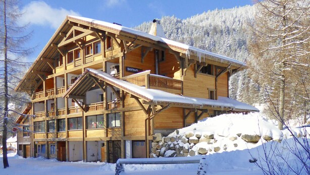 Un chalet en bois à plusieurs étages, doté de balcons, d’un mur en pierre et surplombant un paysage enneigé.