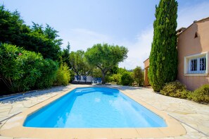Una piscina rectangular con borde de piedra, rodeada de exuberante vegetación y un edificio con una ventana a la derecha.