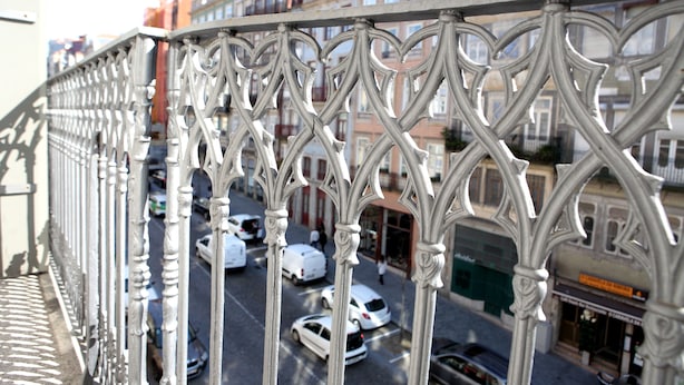 Vista de la calle con barandales de metal decorativos, coches aparcados y edificios con comercios.