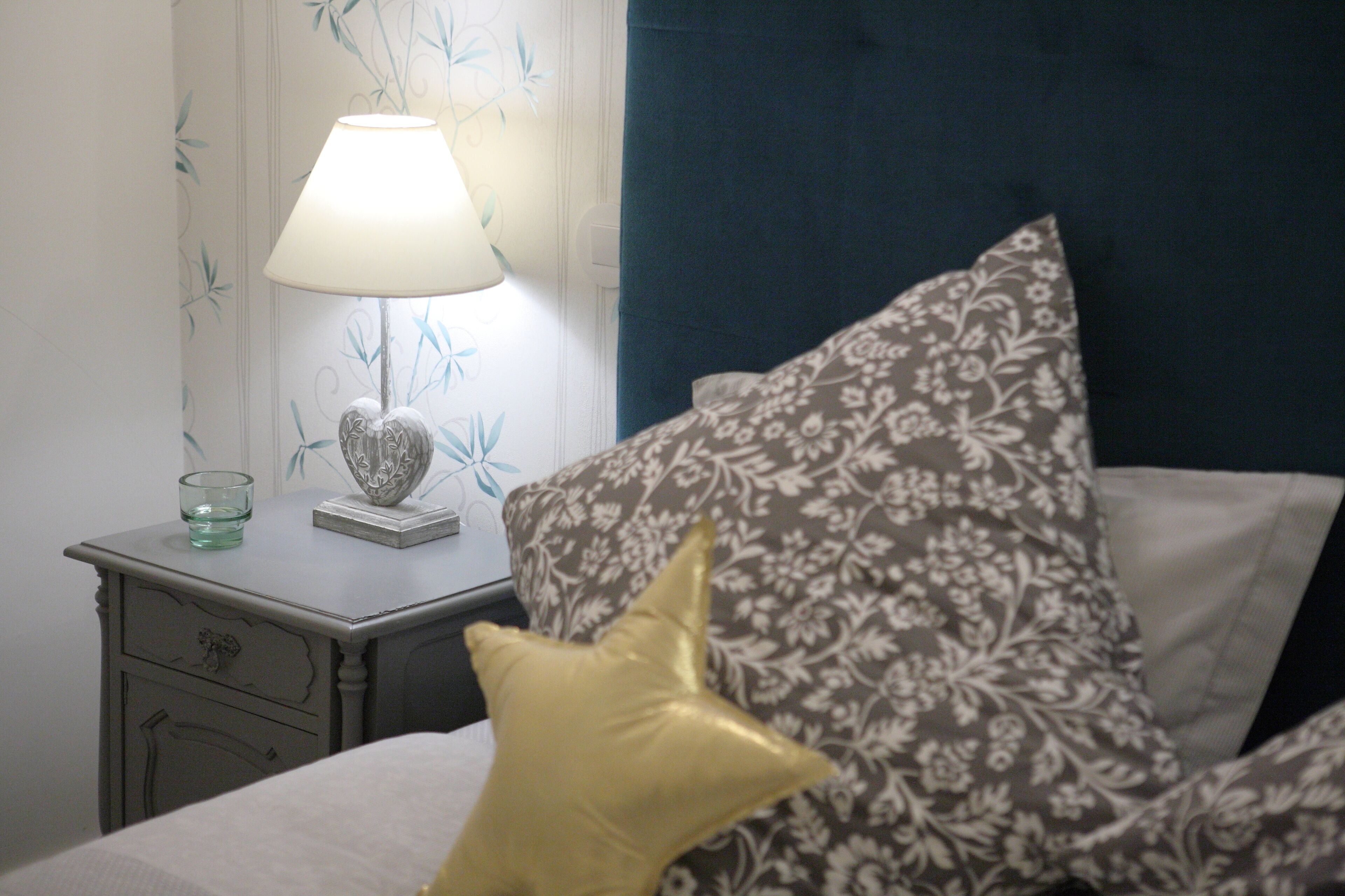 A neatly made bed with a floral pillow and a heart-shaped pillow, a bedside table with a lamp and a glass, and a wall with a leaf pattern.