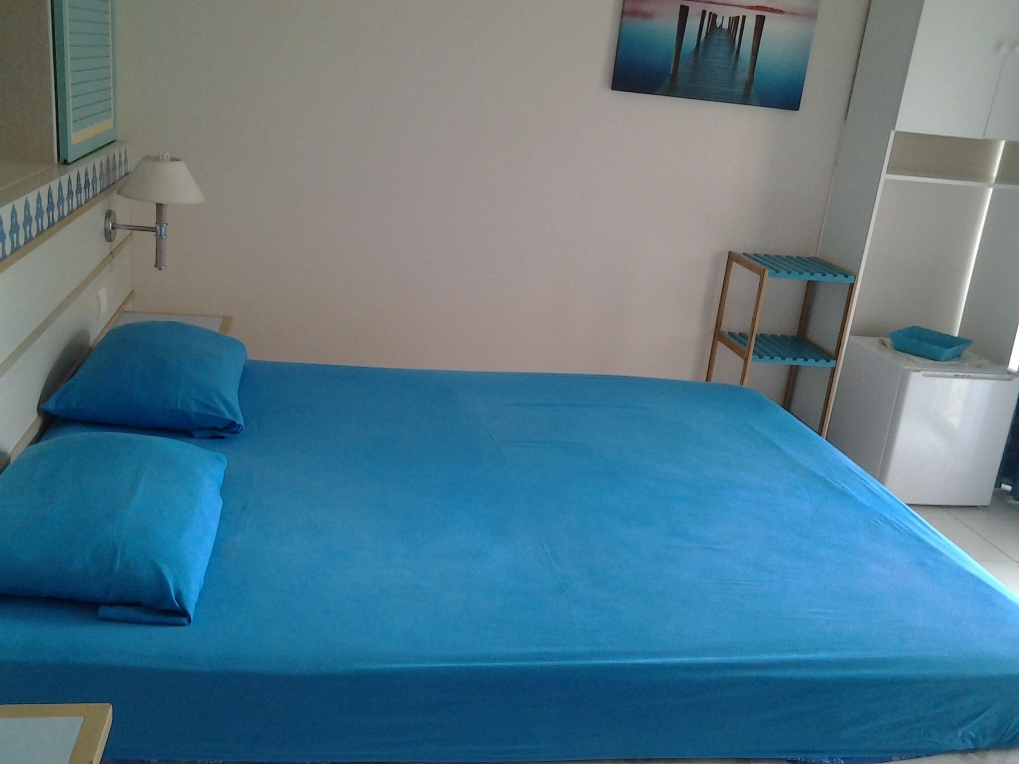 A bed with blue bedding, a wall-mounted lamp, a painting, and a small shelf unit.