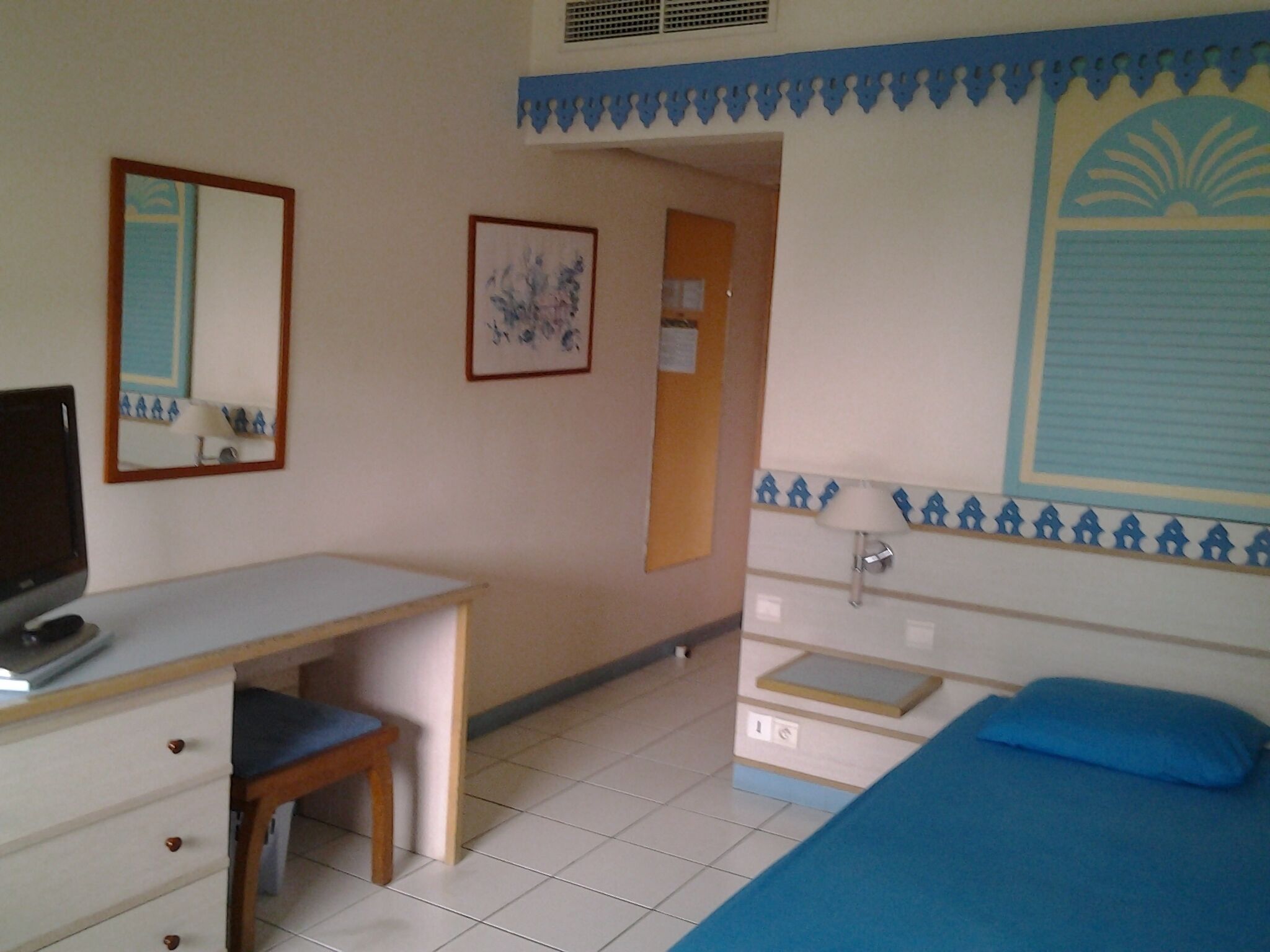 A bedroom with a bed, a desk, a chair, a mirror, and a wall with a decorative pattern.