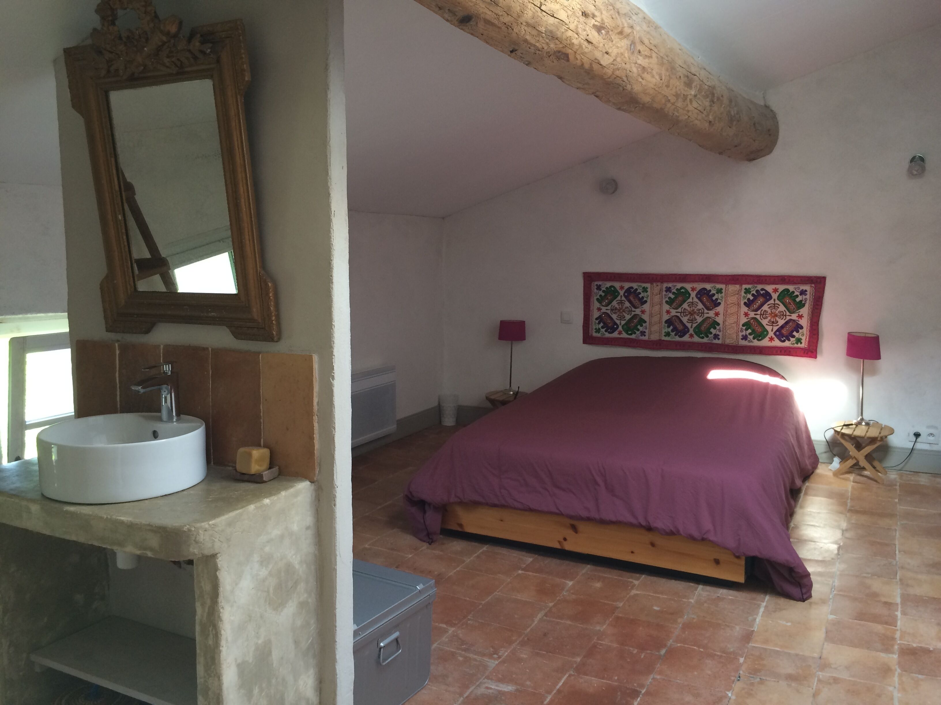 A bedroom with a bed, a bedside table, a lamp, a wall-mounted quilt, and a bathroom with a sink and mirror.