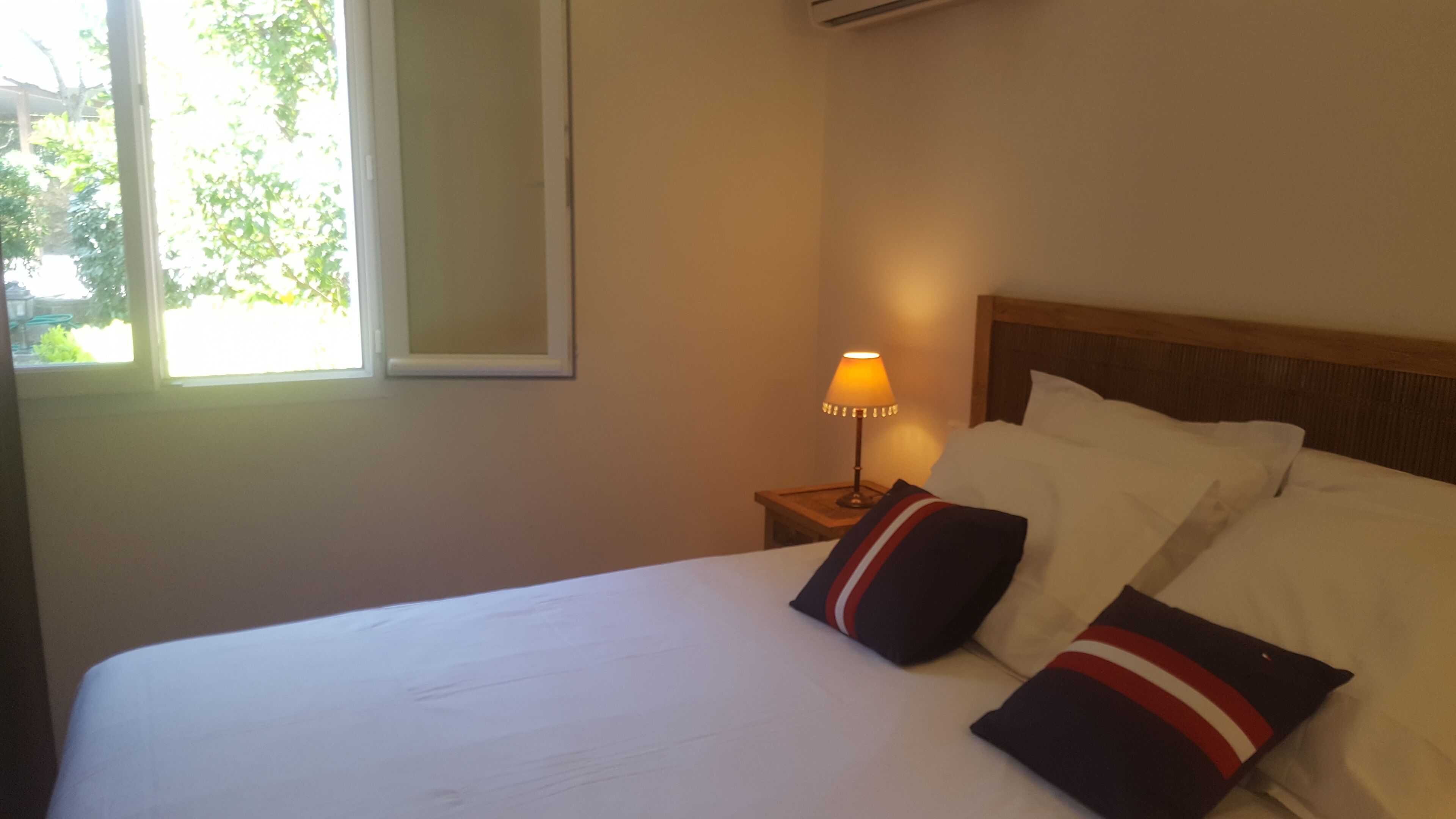 A hotel room with a bed, pillows, a nightstand with a lamp, and a window with a view of greenery.
