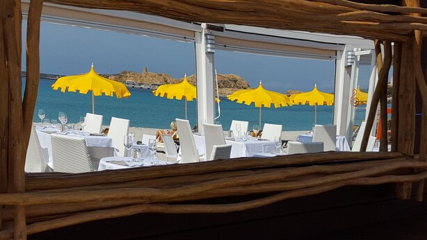 Een restaurant aan het strand met terras, gele parasols en uitzicht op de zee en rotskliffen.